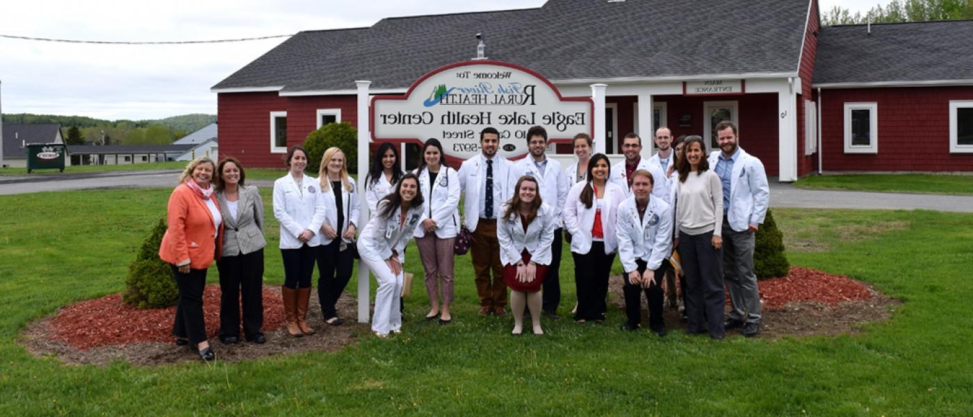 A group of U N E 健康 professions students pose in white coats in front of the sign for Fish River Rural 健康 Center